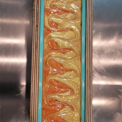 Close-up of freshly poured soap in a mold. The swirled design is in colors of gold and orange. The mold is on a stainless-steel surface.