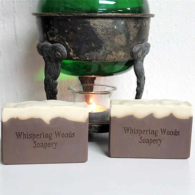 Two rectangular bars of Whispering Woods Soapery soap in a brown and cream color, displayed in front of a vintage warmer with green liquid inside the glass and a lit tealight candle.