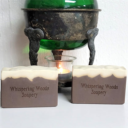 Two rectangular bars of Whispering Woods Soapery soap in a brown and cream color, displayed in front of a vintage warmer with green liquid inside the glass and a lit tealight candle.