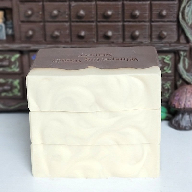 Three stacked rectangular bars of soap with a dark brown color on the bottom and cream on top. Stamped 'Whispering Woods Soapery.' The stack is set against a blurred background of a small apothecary cabinet.