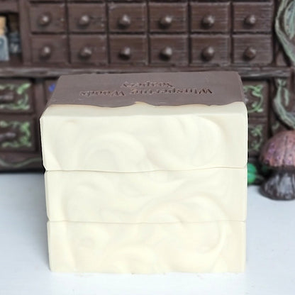 Three stacked rectangular bars of soap with a dark brown color on the bottom and cream on top. Stamped 'Whispering Woods Soapery.' The stack is set against a blurred background of a small apothecary cabinet.