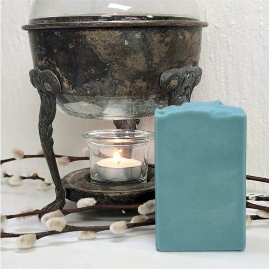 A greenish-blue bar of soap sits on a white surface in front of a vintage warmer with a lit tealight candle.