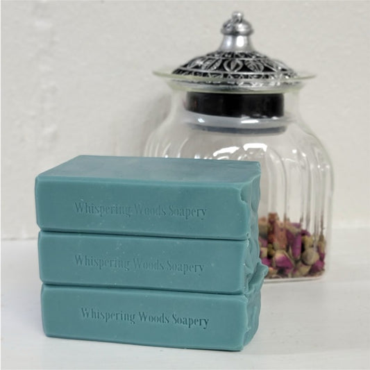 Three stacked bars of Whispering Woods Soapery soap in a dusty teal-blue color, placed in front of a clear glass jar with a silver lid containing dried rose petals.