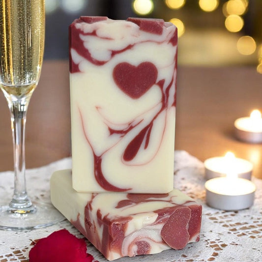 Two off-white bars of soap with cranberry-colored swirls and small heart embed on a candlelit table with a glass of champagne