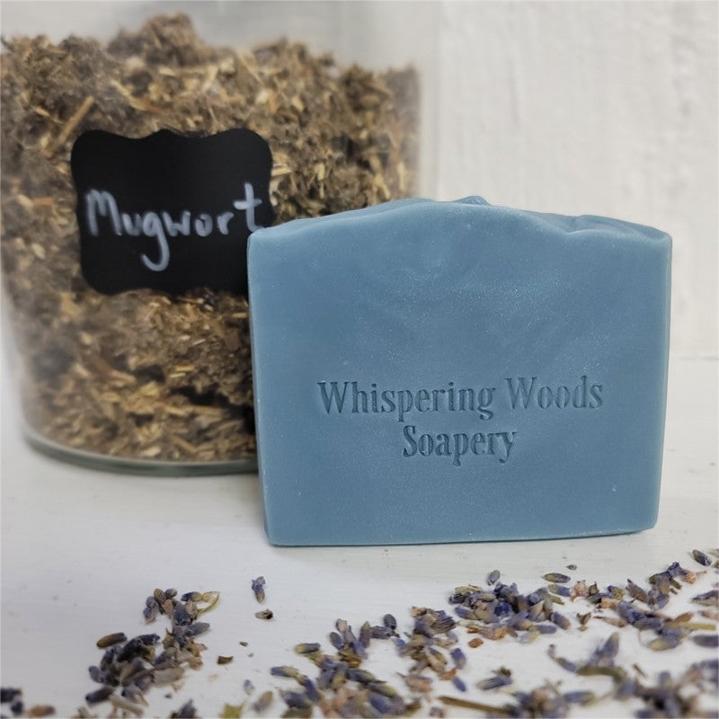 A dusky-blue bar of soap stamped 'Whispering Woods Soapery' in front of a large glass jar filled with the herb mugwort. There are lavender buds strewn around on the white surface.