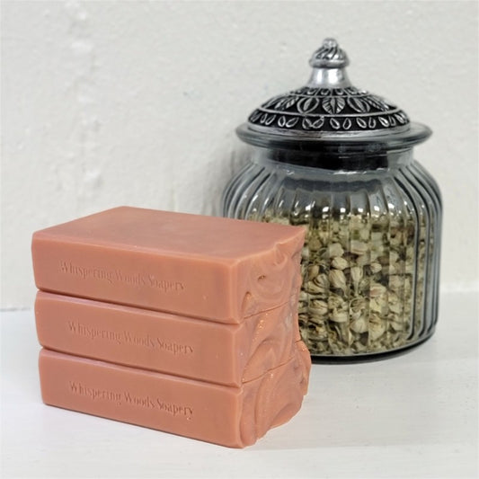 Three stacked rectangular bars of Whispering Woods Soapery soap in a dusty terracotta pink color, positioned in front of a ribbed clear glass jar with a silver-toned lid containing dried botanicals, set against a white textured wall.