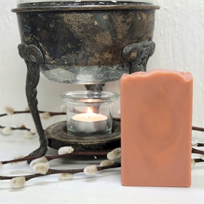 A rectangular peachy-pink-colored soap bar stands next to a vintage warmer holding a lit tealight candle against a white wall. There are decorative catkins in the foreground.