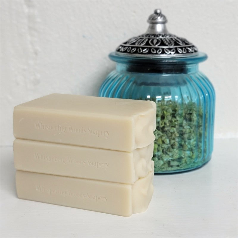 Three stacked tan-colored soap bars with stamped text reading 'Whispering Woods Soapery,' next to a blue ribbed glass jar filled with dried herbs, set against a white textured wall.