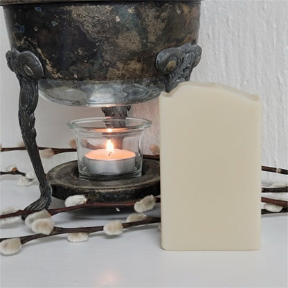 A light tan rectangular bar of soap stands next to a lit tealight candle in a clear glass holder in a vintage warmer. There are decorative pussy willow branches in the scene.