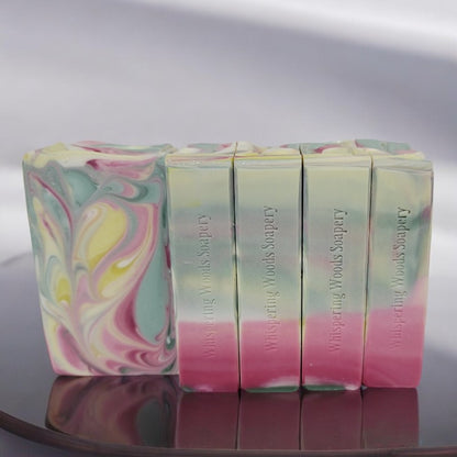 Five bars of light green, white, yellow, and pink swirled soap bars on a reflective surface with a white background