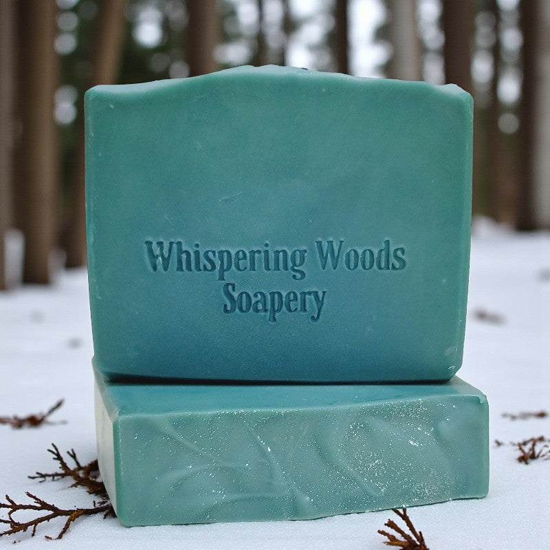Two bars of teal soap with 'Whispering Woods Soapery' branding on a snowy background