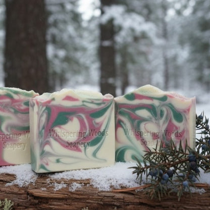 Three bars of soap with a marbled design on a log with a snowy forest background, featuring the brand 'Whispering Woods Soapery'