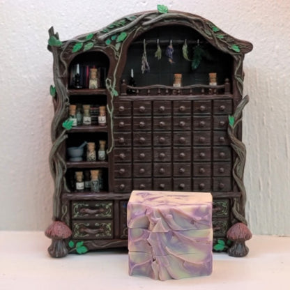 A miniature apothecary cabinet display with many small drawers and shelves holding miniature bottles and jars against a textured white wall. In front of the cabinet is a stacked set of rectangular soap bars with a swirled lavender and cream pattern.
