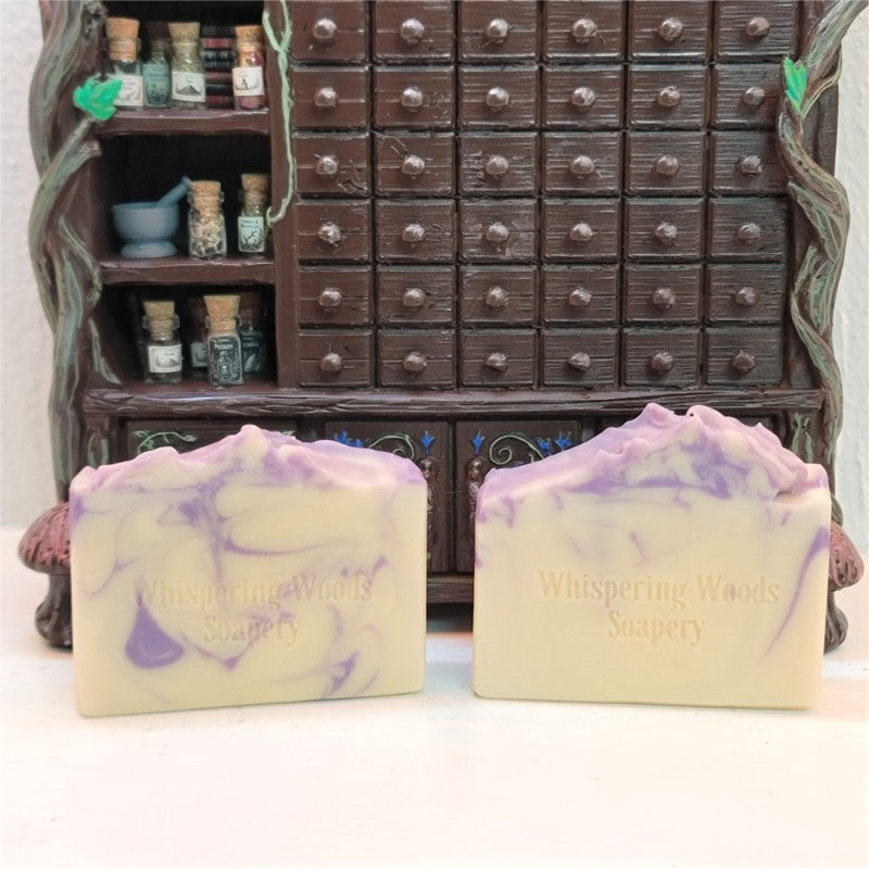 Two handmade rectangular bars of goat milk soap, cream with lavender-colored swirls, stamped with 'Whispering Woods Soapery,' displayed in front of a miniature dark brown apothecary cabinet.