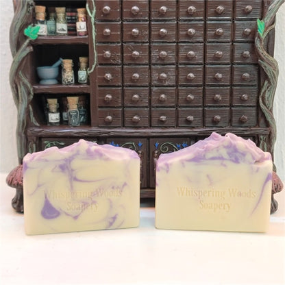 Two handmade rectangular bars of goat milk soap, cream with lavender-colored swirls, stamped with 'Whispering Woods Soapery,' displayed in front of a miniature dark brown apothecary cabinet.