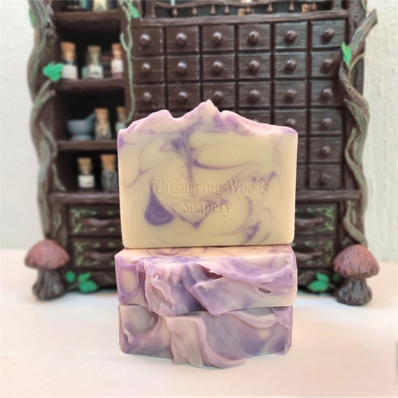 Three handmade soap bars, cream with lavender swirls. The top bar has 'Whispering Woods Soapery' stamped in it. A decorative dark wood apothecary-style cabinet is blurred in the background.