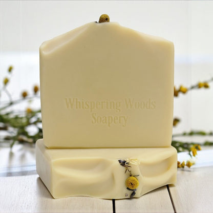 Bars of soap with 'Whispering Woods Soapery' branding on a wooden surface with blurred natural background