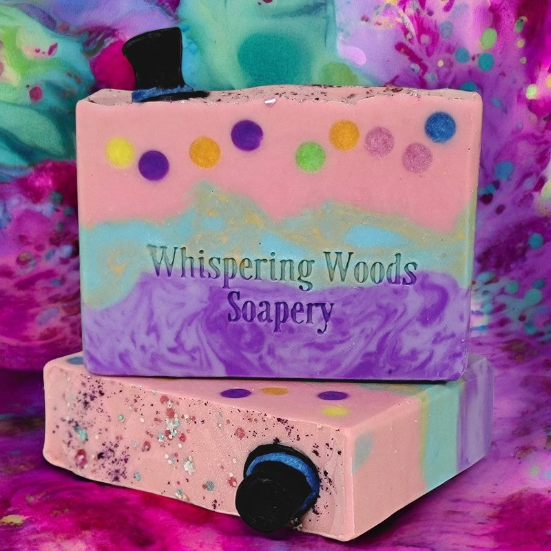 Two bars of pink, teal and purple soap, with glitter and small black top hats on top, with a multicolored background