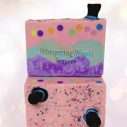 Three bars of pink, teal and purple soap, with glitter and small black top hats on top, with soft lighting in the background