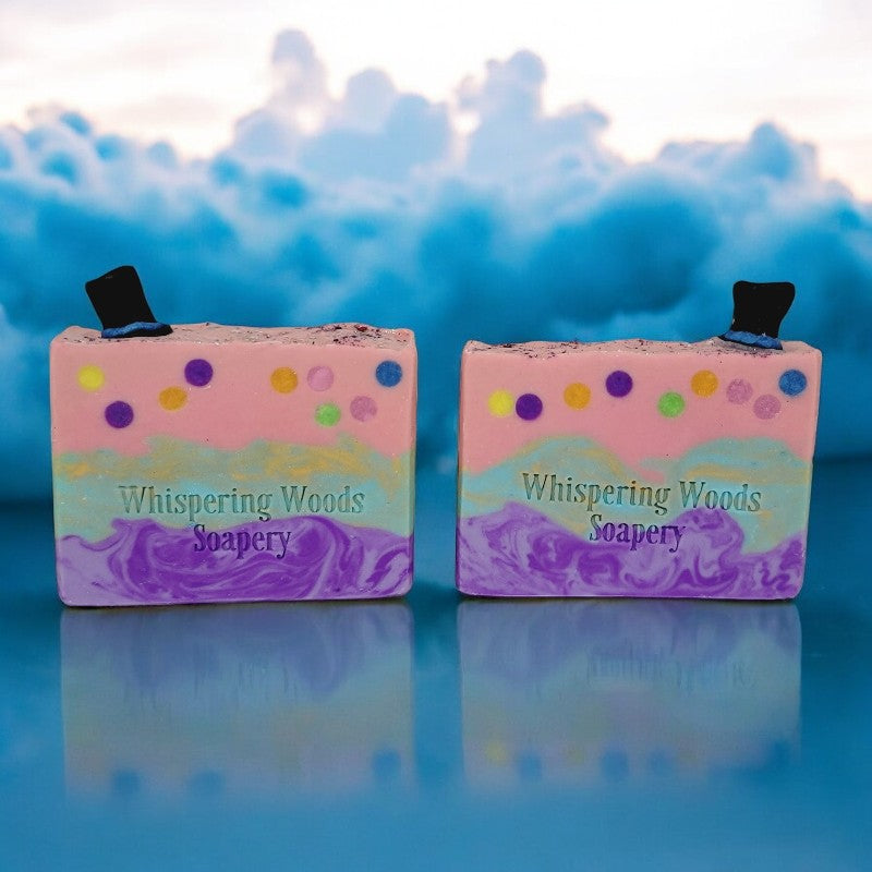 Two bars of pink, teal and purple soap, with glitter and small black top hats on top, on a blue reflective surface with clouds in the background