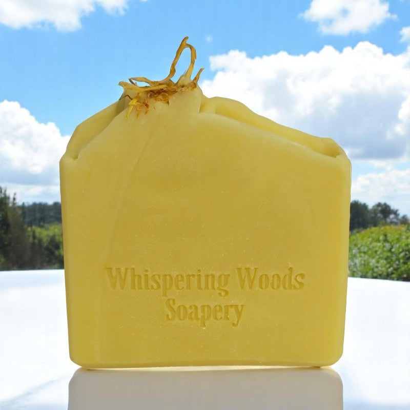 A bar of yellow soap with dried calendula on top on a white marble table. There is blue sky and white clouds in the background.