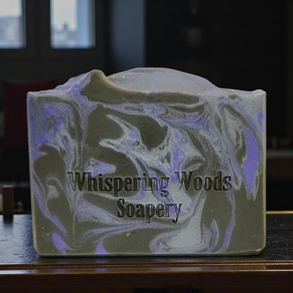 Silver, purple and black swirled soap bar with 'Whispering Woods Soapery' branding on a dark surface