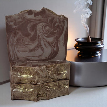 Three bars of brown-swirled soap with sparkly gold tops on a counter with a cone of incense burning beside them