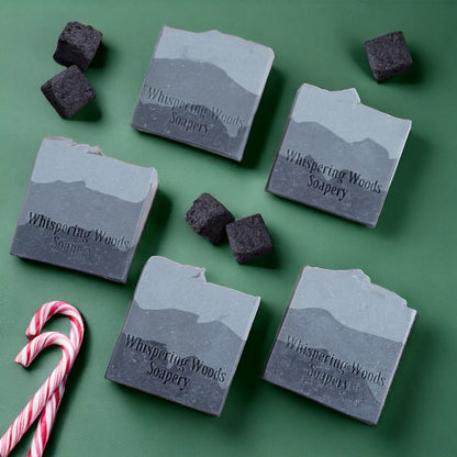 Five bars of soap lay flat on a green surface. The soap has three layers of charcoal gray, with the darkest on the bottom and lightest at the top. There are a couple candy canes and lumps of coal scattered about.