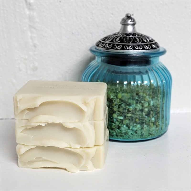 Three stacked bars of creamy off-white handmade soap in front of a blue ribbed glass jar with a silver metal lid, set against a white textured wall.