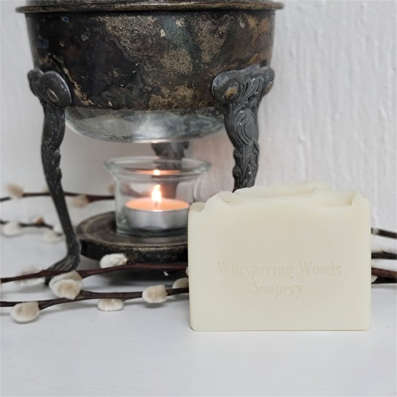 An ivory-colored soap bar, stamped 'Whispering Woods Soapery,' in front of a vintage warmer with lit tealight candle on a white surface.