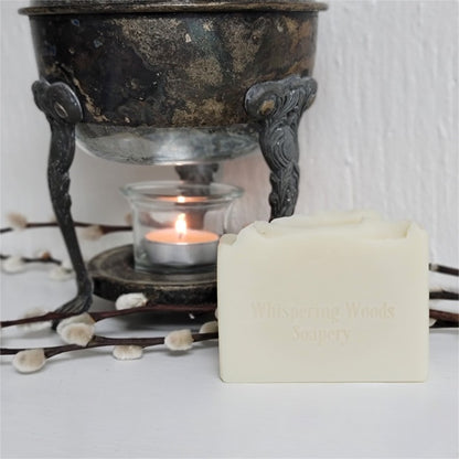 An ivory-colored soap bar, stamped 'Whispering Woods Soapery,' in front of a vintage warmer with lit tealight candle on a white surface.
