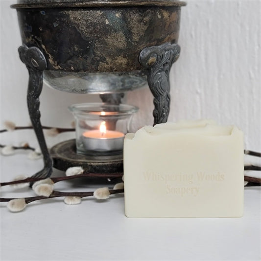 An ivory-colored soap bar, stamped 'Whispering Woods Soapery,' in front of a vintage warmer with lit tealight candle on a white surface.