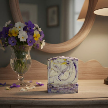 White soap with purple and yellow swirls on a wooden surface next to a vase of violets and a mirror