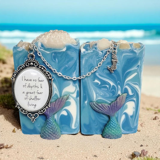 Two bars of soap with gorgeous blue, green and white swirls on a sandy beach with the ocean in the background. A locket with the words "I have no fear of depths & a great fear of shallow living" drapes across the soap bars.