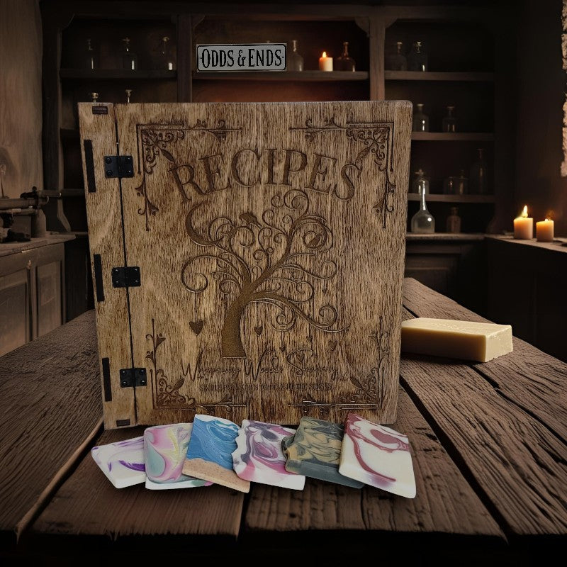Whispering Woods Soapery's recipe book and some soap bundle ends on a wood table with a shelf of bottles and some candles in the background