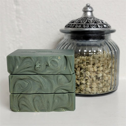 Three stacked rectangular bars of green soap with textured tops and gold sparkle, displayed next to a glass jar filled with dried botanicals on a white surface against a textured white wall.