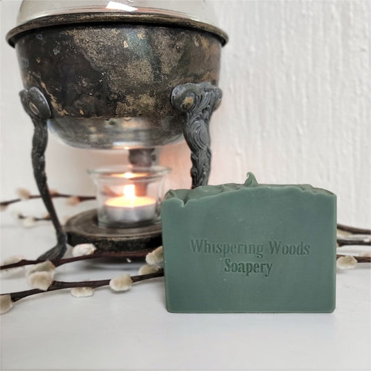 A rectangular bar of dark green soap from Whispering Woods Soapery sits on a white surface in front of a vintage metal warmer with a lit tealight candle. Willow branches are arranged around the scene.
