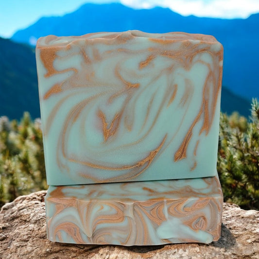 Two bluish-green and copper swirled soap bars sit on a rock with a mountain in the background