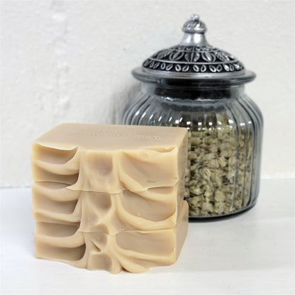 A handmade rectangular stack of soap  bars in a warm beige tone with textured tops, displayed in front of a ribbed clear glass jar with a silver-toned lid filled with dried botanicals, set against a white textured background.