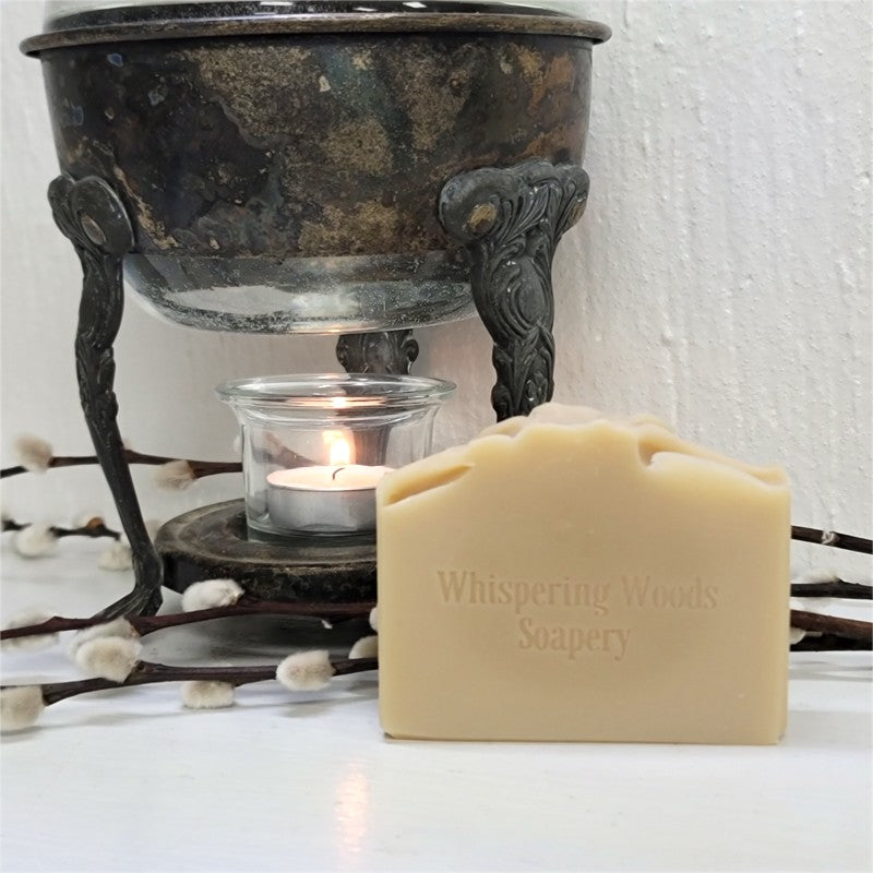 A rectangular bar of Whispering Woods Soapery soap in a warm cream color, placed in front of a vintage warmer with a lit tealight candle. The scene is decorated with willow branches.
