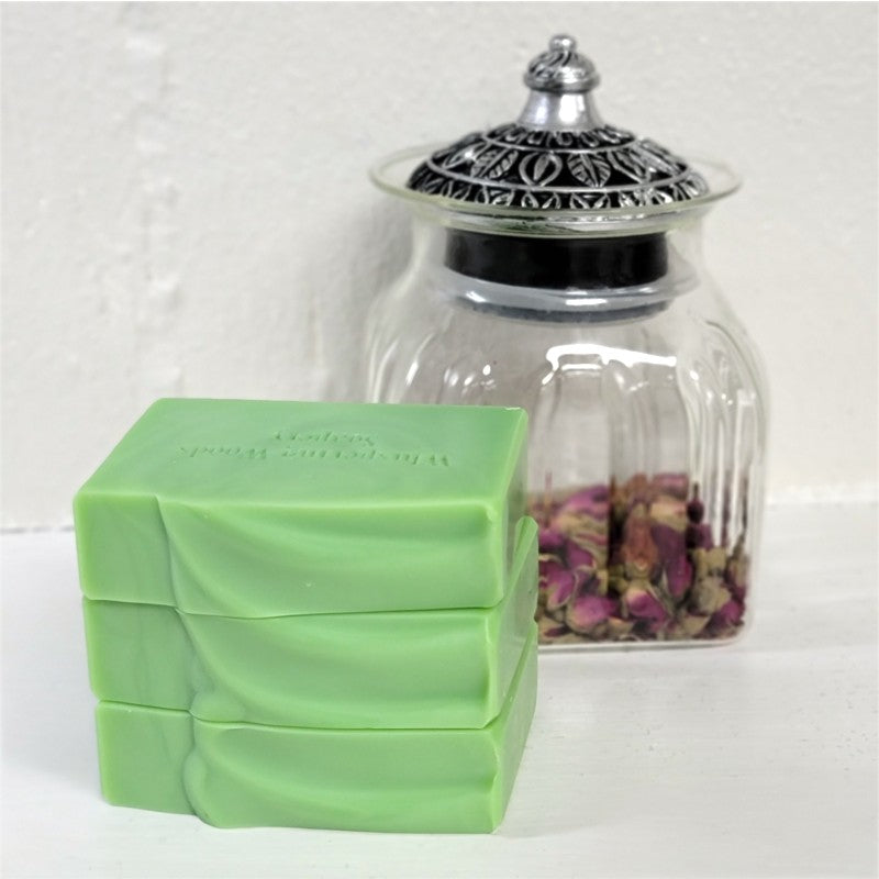 Three stacked light green rectangular bars of soap in front of a clear glass jar with a silver lid, containing dried pink flowers, on a white surface against a white wall.