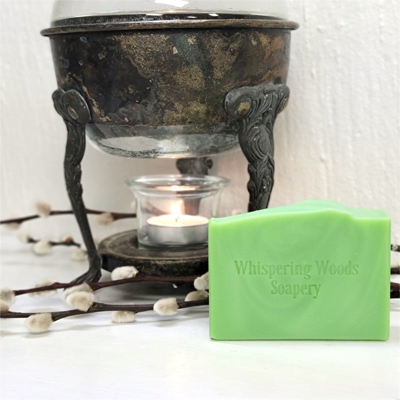 A rectangular bar of light green soap from Whispering Woods Soapery sits on a white surface in front of a dark metal warmer with a lit tealight candle. Willow branches are scattered around the base.