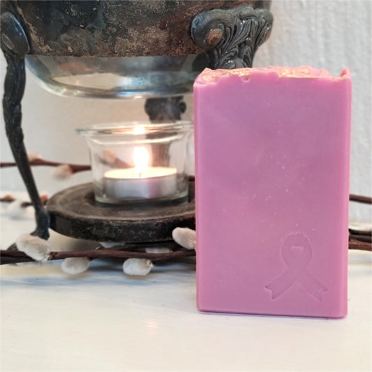 A rectangular pink bar of soap with a breast cancer ribbon stamped in the right bottom corner, on a white surface with a lit tealight candle.