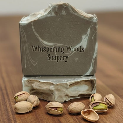 Two bars of soap with the branding 'Whispering Woods Soapery' sit on a wood surface with some pistachios scattered about