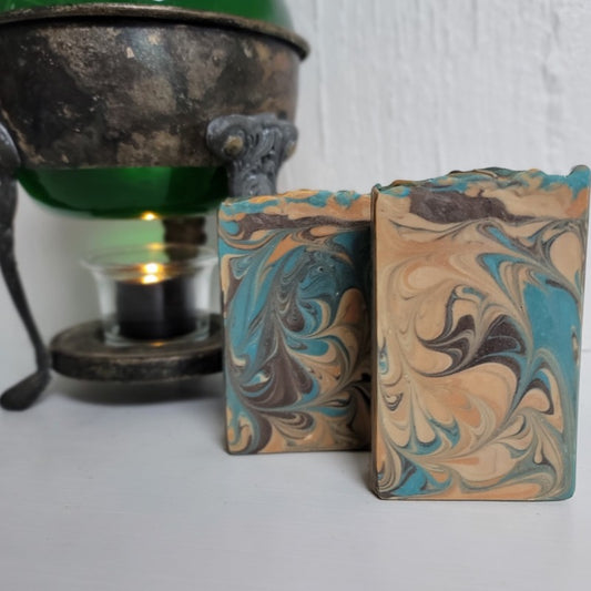 Two rectangular soap bars with teal, brown, orange and champagne swirls stand in front of a vintage warmer with a lit tealight candle.