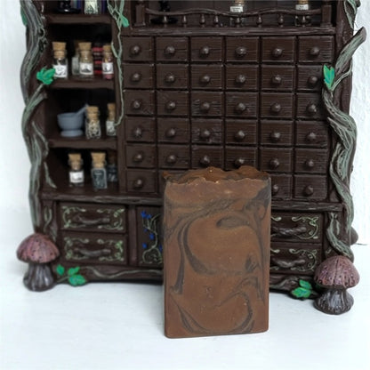 A single rectangular bar of medium brown soap with darker swirls in front of a miniature apothecary cabinet on a white surface.