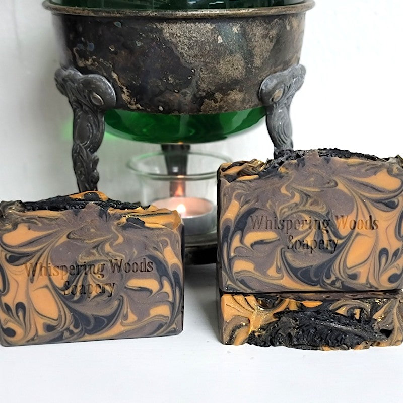 Three handmade soap bars with a marbled swirl pattern of black, brown, and burnt orange, displayed in front of a vintage warmer with a lit candle. The soap is stamped with the brand name 'Whispering Woods Soapery' and features a black quill feather made of soap dough on top.