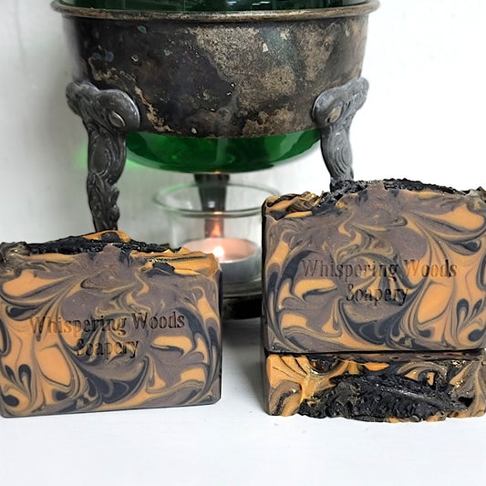 Three handmade soap bars with a marbled swirl pattern of black, brown, and burnt orange, displayed in front of a vintage warmer with a lit candle. The soap is stamped with the brand name 'Whispering Woods Soapery' and features a black quill feather made of soap dough on top.