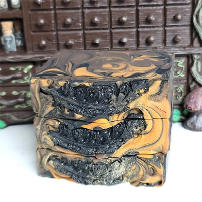 Three stacked rectangular bars of marbled soap with a black, brown and burnt orange swirl pattern, set against a dark wooden apothecary cabinet background. The tops have black soap dough quill feathers and gold sparkle.