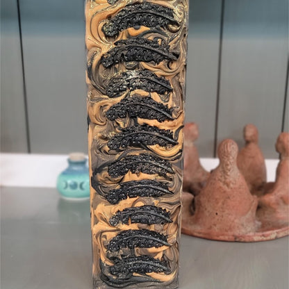 A soap loaf in hues of brown, orange and gold stands vertically on a gray surface. There are black soap-dough quill feathers along the top and gold sparkle. Pottery pieces are in the background.